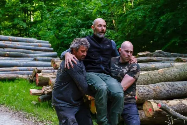 Zwei Personen tragen einen dritten Mann zwischen sich über einen Holzstapel